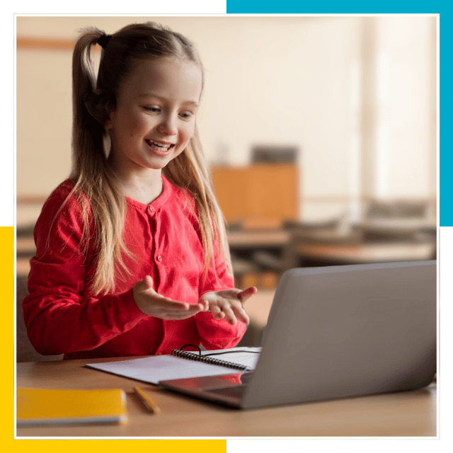 A young girl wearing a red sweater sits at a desk with a laptop, notebook, and pencil, participating in an online class in a classroom setting.