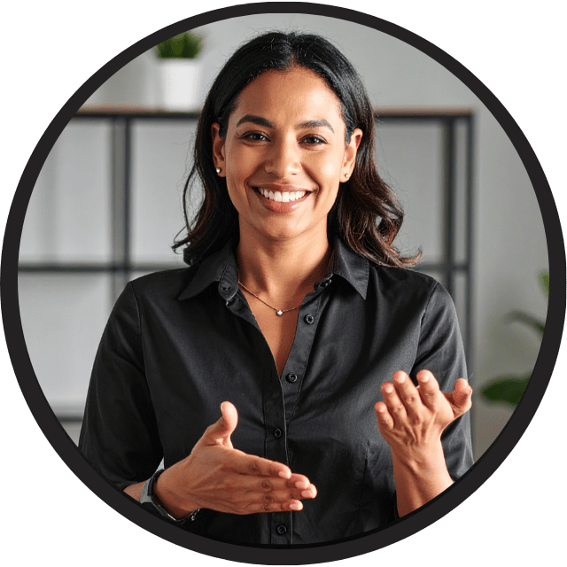 A person with shoulder-length dark hair is wearing a black button-up shirt and a necklace, standing indoors in front of a shelf with a plant and other items. The individual is gesturing with both hands, suggesting they may be communicating or explaining something, possibly using sign language.