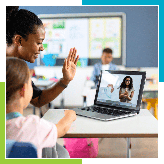 A teacher and a young student sit together at a classroom desk, interacting with a laptop. On the laptop screen, a person is communicating using sign language during a video call. The classroom setting is bright and colorful, with educational materials visible in the background. This scene highlights the use of technology to support remote learning and sign language communication in an educational environment.